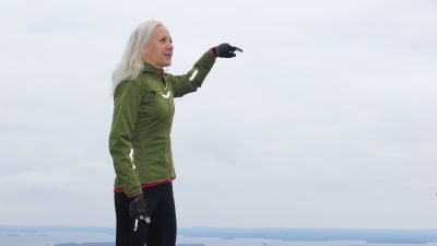Kaisa Mäkäräinen står högt uppe på ett berg och pekar ut över Koli nationalpark