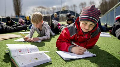 Elever i S:t Karins svenskspråkiga skola ligger på en gräsmatta och skriver i häften.