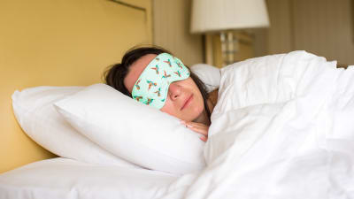 Kvinna ligger under täcket med en sovmask över ögonen.