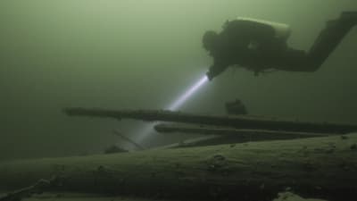En dykare som lyser med en ficklampa på ett gammalt skeppsvrak på havsbotten.
