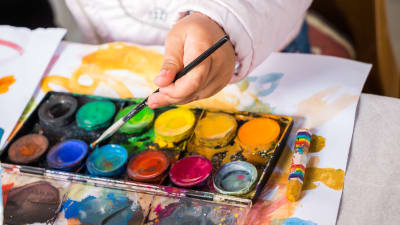 Olika färger av vattenfärg och handen av ett barn som håller i en pensel.