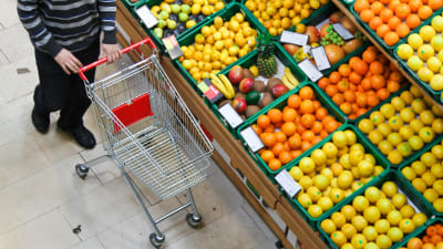 En person skuffar en kundvagn framför ett stort sortiment av bland annat citrusfrukter.