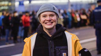 En leende kvinna i mössa och gul regnrock i förgrunden. I bakgrunden människor på en gata.