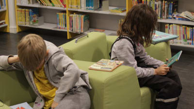Två stycken barn sitter och läser i ett bibliotek.