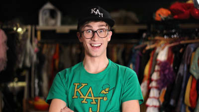 Christoffer Strandberg utklädd till medlemmarna i humorgruppen KAJ. Han bär svart keps och grön t-skjorta som det står KAJ på.