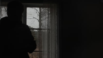En anonym person står vid ett fönster. Utanför syns bara träd.