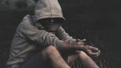 En ung man i grå huvtröja sitter i gräset och injicerar en spruta in i sin arm.