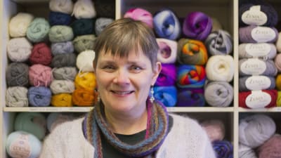 En kvinna med kort ljusbrunt hår står framför en hylla fylld med garn.