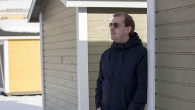 En man som står och lutar sig mot en husvägg. Han har på sig solglasögon.
