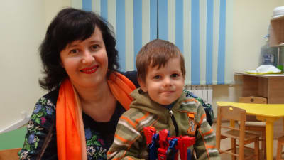 Svetlana i orange scark och blå klänning med sin son i famnen. Sonen håller en transformersfigur i handen