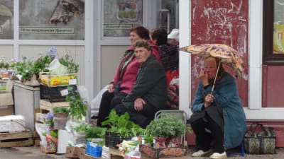 Många säljer en del av sin skörd på marknadsplatser. tre äldre kvinnor säljer olika grödor. En sitter under ett paraply.