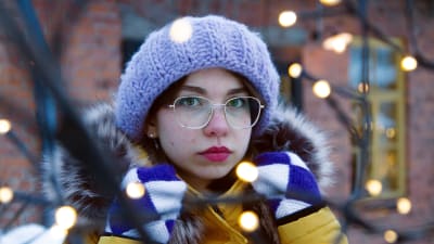 20-vuotias sosiaalisen median sisällöntuottaja Saara Strum seisoo oksiin aseteltujen jouluvalojen takana. Hän pitää molemmilla käsillä kiinni talvitakkinsa hupun kauluksista ja katsoo suoraan kameraan.
