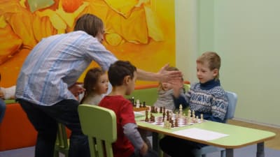 En schacklärare berömmer barn, fyra barn sitter vid ett bord. 