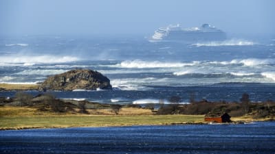 En bild på ett kryssningsfartyg i kraftig storm. I förgrunden syns land och hav.