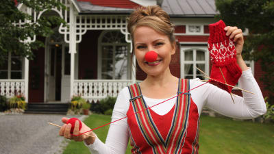 Tv-programmet Strömsös hantverksexpert Lee Esselström står framför Strömsövillan med en röd clownnäsa och håller en halvfärdig röd yllestrumpa. Yllestrumpan är hennes design för Näsdagen 2020.