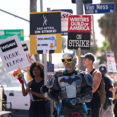 En man i Batman-klädsel strejkar, i bakgrunden syns människor som håller upp plakat om strejken.