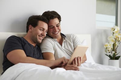 Två leende män ligger i en säng och tittar på en pekskärm