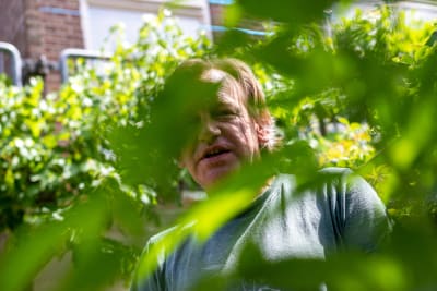 En man ute i sin trädgård fotad genom en växt vars blad syns suddiga i förgrunden.