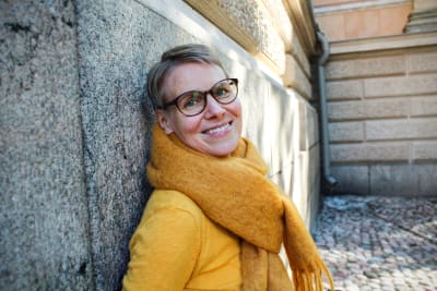 En yngre brett leende, glasögonprydd kvinna klädd i gul tröja och gul halsduk lutar sig mot en husgrund 