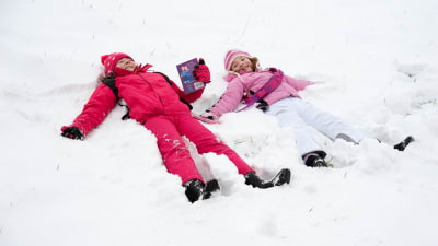 Två barn ligger i en snöhög på rygg. De skrattar.