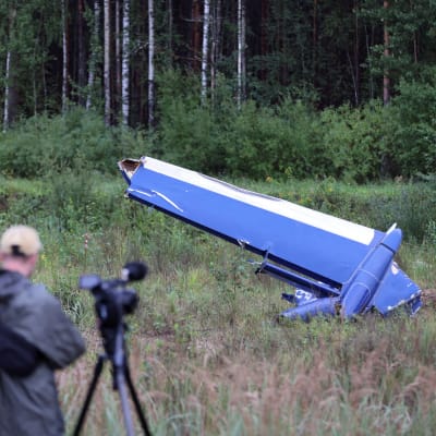 En man fotograferar en flygplansvinge som ligger på marken en bit framför honom en bit från ett skogsbryn.