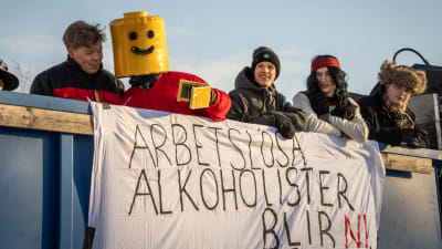 Utklädda aiturienter står på ett lastbilsflak. Framför dem hänger ett lakan där det står "Arbetslösa alkoholister blir ni".