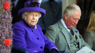 Drottning Elizabeth sitter bredvid prins Charles. Hon iklädd en lila rock med tillhörande hatt och han med en grå kostym. 