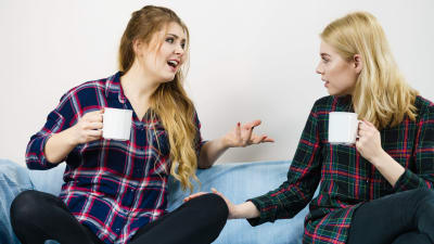 Två kvinnor med kaffekoppar i soffa, den ena gestikulerar och pratar, den andra sitter tyst