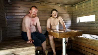 Två män med bar överkropp sitter bredvid varandra på en lave i en träbastu.