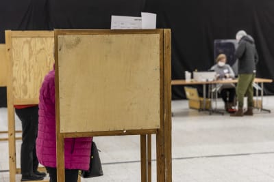 I förgrunden kvinna i rosa ytterock som står i valbås för att avlägga sin röst. I bakgrunden valfunktionär som stämplar valsedel.