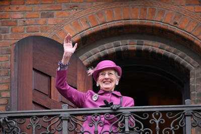 Drottning Margrethe iklädd lila hatt och kappa vinkar glatt på en balkong.