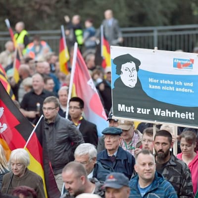Ryhmä mielenosoittajia seisoo lippujen ja plakaattien kanssa. Yhdessä julisteessa on Martti Lutherin kuva ja tekstinä: "Tässä seison enkä muuta voi" ja "Rohkeutta totuuteen!" Lisäksi on Angela Merkelin kuva tekstillä "Compact" ja "Mutti multikulti".