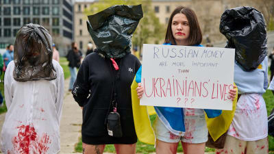 Kvinnor med blodiga kläder och svarta sopsäckar på huvudet demonstrerar i Berlin mot Tysklands ovilja att bojkotta rysk olja och gas. En av kvinnorna är iklädd en ukrainsk flagga och håller upp en skylt med texten "Ryska pengar är viktigare än ukrainska liv?". 