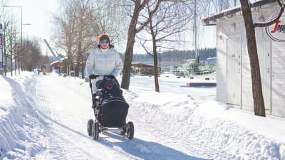 Kvinna med barnvagn går längs en plogad väg i solsken.