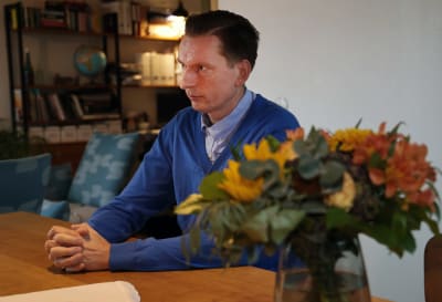 Profilbild av Henry Johansson som sitter vid ett bord med händerna knäppta. Han bär en blå tröja och kragskjorta.
