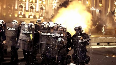 Kravallpoliser i full mundering står framför parlamentet i Belgrad. I bakgrunden syns en explosion av något slag.
