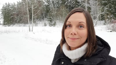 Saija Westerlund-Cook, en dam med rakt mörkt hår, står i ett snöigt åker- och skogslandskap.