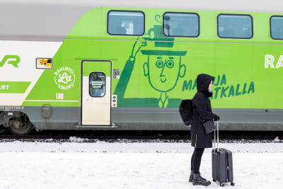 En kvinna med liten dragväska står på en tågperrong med ett tåg bakom sig. 