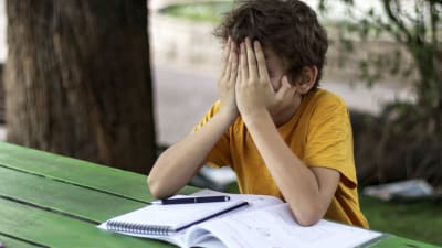 Ett barn sitter vid ett bord med skolböcker framför sig. Barnet håller sitt ansikte i sina händer och ser ledset ut.