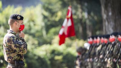 En soldat från Schweiz står i förgrunden med munskydd på. I bakgrunden fler soldater samt Schweiz flagga.