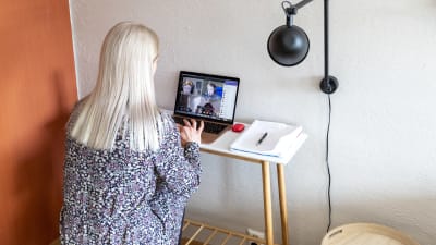 Läraren Susanne Ojaniemi undervisar sin klass via en dator på distans i sit them. Hon har datorn på ett skrivbord av trä och svart lampa syns bredvid bordet där Ojaniemi sitter.