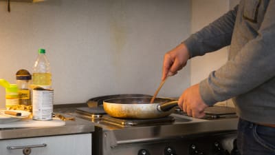 En man står vid en spis och lagar mat i en stekpanna. På köksbänken står öppnade konserver och matolja.