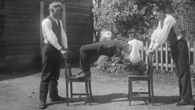 En man böjer sig uppåt mellan två stolar med hjälp av bara benen och huvudet, inte händerna. Bilden är tagen 1931 i Närpes.