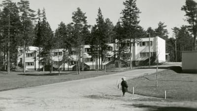 Liten pojke står på en sandväg, i bakgrunden ett nybyggt vitt tvåvåningsradhus. Bilden är svartvit och tagen på 1950-talet.