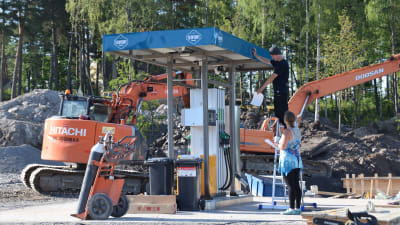 En ny bränslestation installeras på ett byggområde. Två personer står och inspekterar stationen. I bakgrunden syns en orange grävskopa.