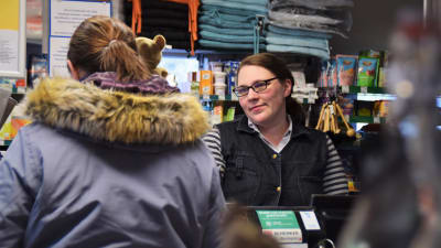En kvinna sitter i en kassa i en liten livsmedelsbutik, i sällskap av en kund som har ryggen mot kameran.