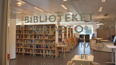 Texten Bibliotek Kirjasto på en glasdörr med många hyllor med böcker i bakgrunden.