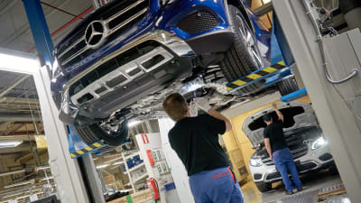 Bilbyggare vid Nystads bilfabrik bygger ihop en mörkblå bil som hänger från taket.