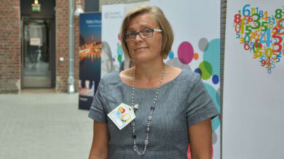 Professor Eva Österbacka i en grå klänning framför planscher och en teglad vägg.