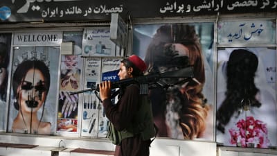 En talibansoldat bär på ett gevär. Han går förbi en affär där ansikten på kvinnor på raklamaffischer målats över.